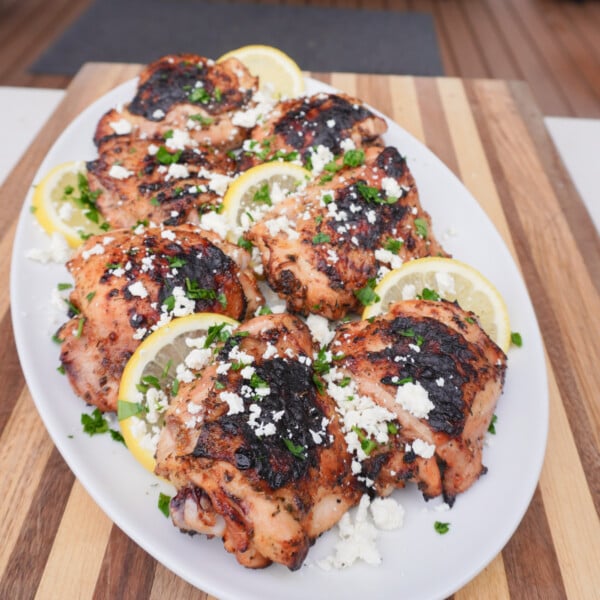 Grilled Greek Lemon Chicken plated on a white platter with sliced lemons, feta cheese and chopped parsley.